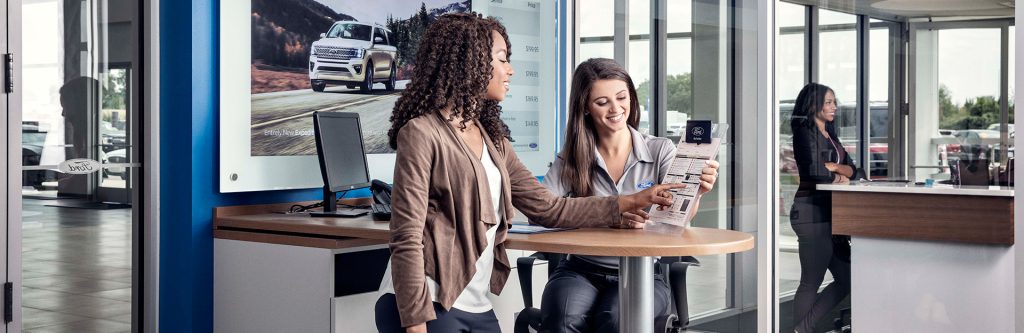 A female customer with curly hair points at a clipboard held by a smiling dealership employee inside a modern Ford car dealership showroom.